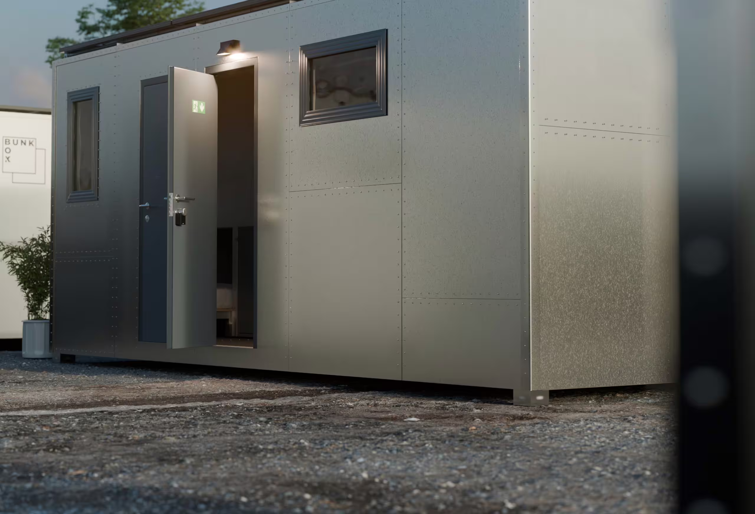 A BunkBox module on gravel at dusk with its personnel door open and the interior lit, an emergency exit sign above the doorway and a small window alongside