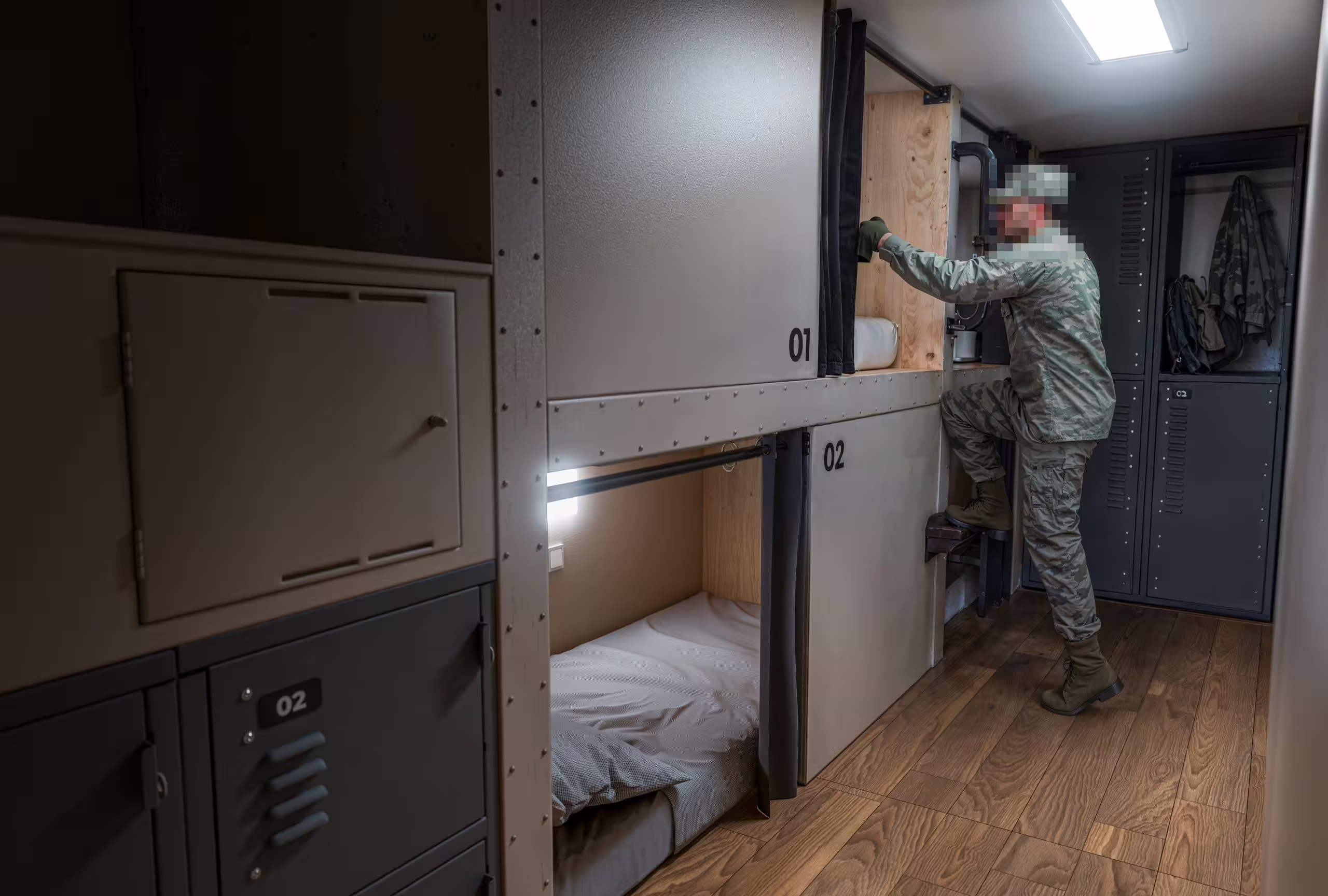 A soldier stepping up to the upper berth inside a BunkBox module, berths numbered 01 and 02 with the lower one made up, ventilated steel lockers and a hung jacket alongside