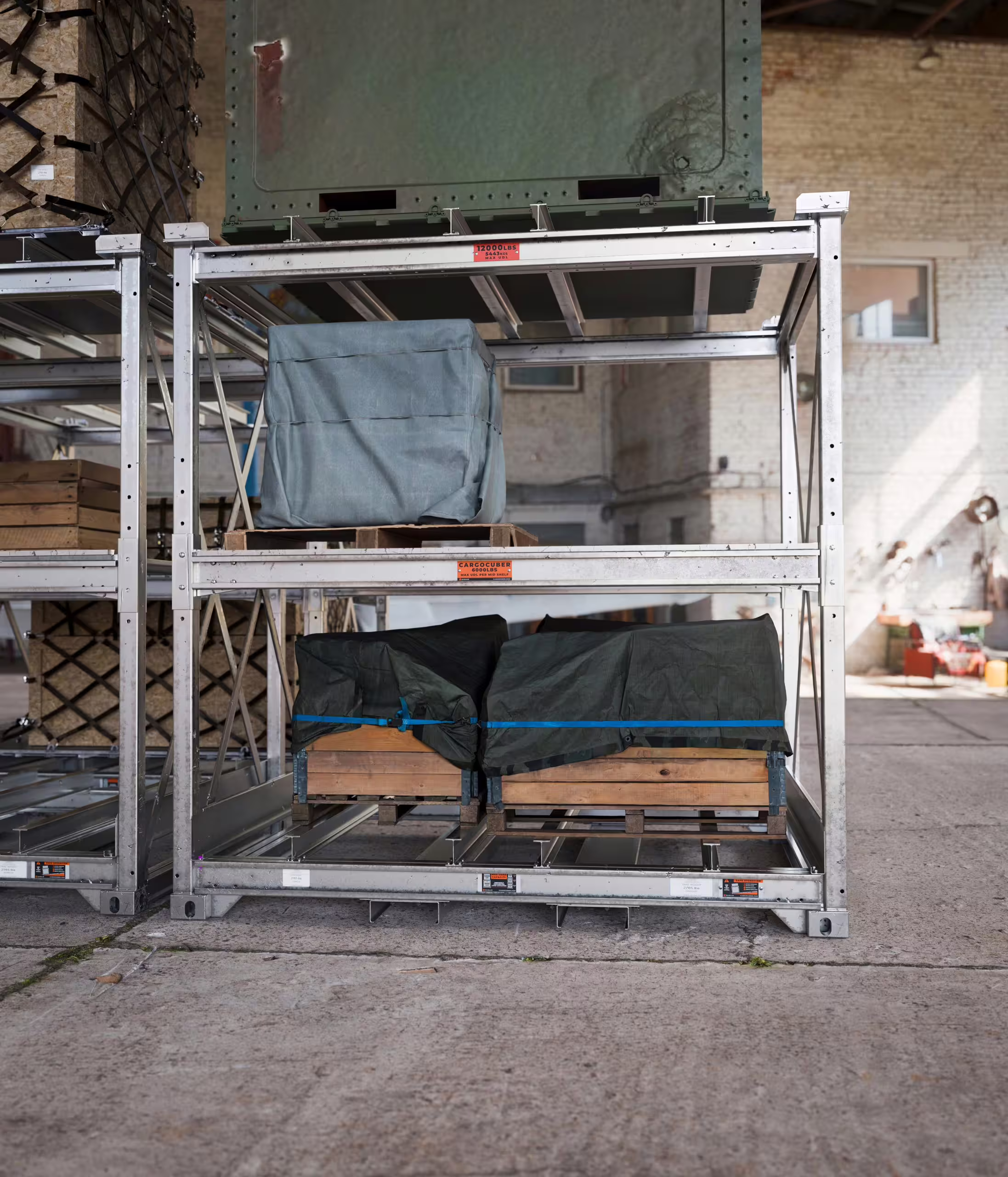 CargoCuber frames standing outdoors against a wall, carrying tarpaulin-covered and strapped loads on timber bearers, with the orange load-rating plates visible on the beams