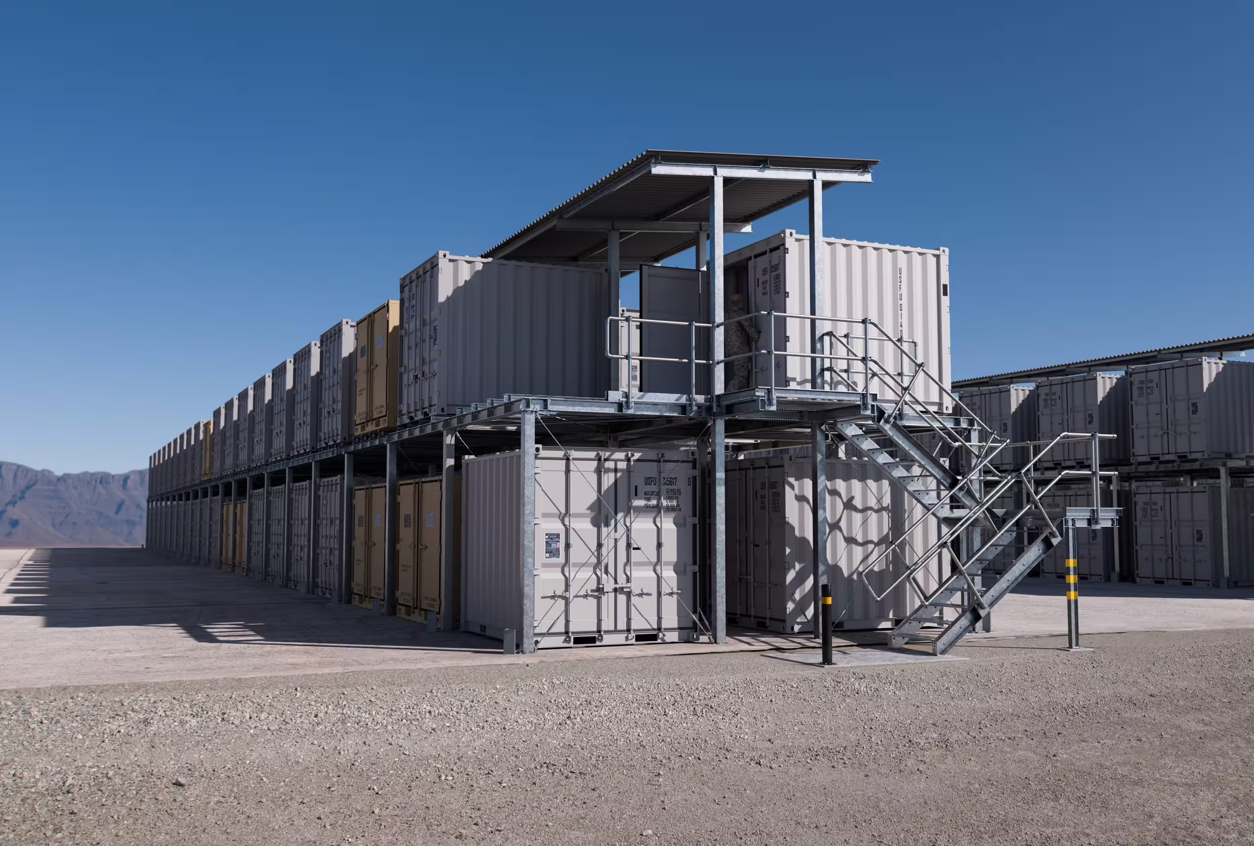 A two-level Universal CargoRack run under a monopitch roof on gravel, containers racked on both levels, an external access stair and landing at the near end