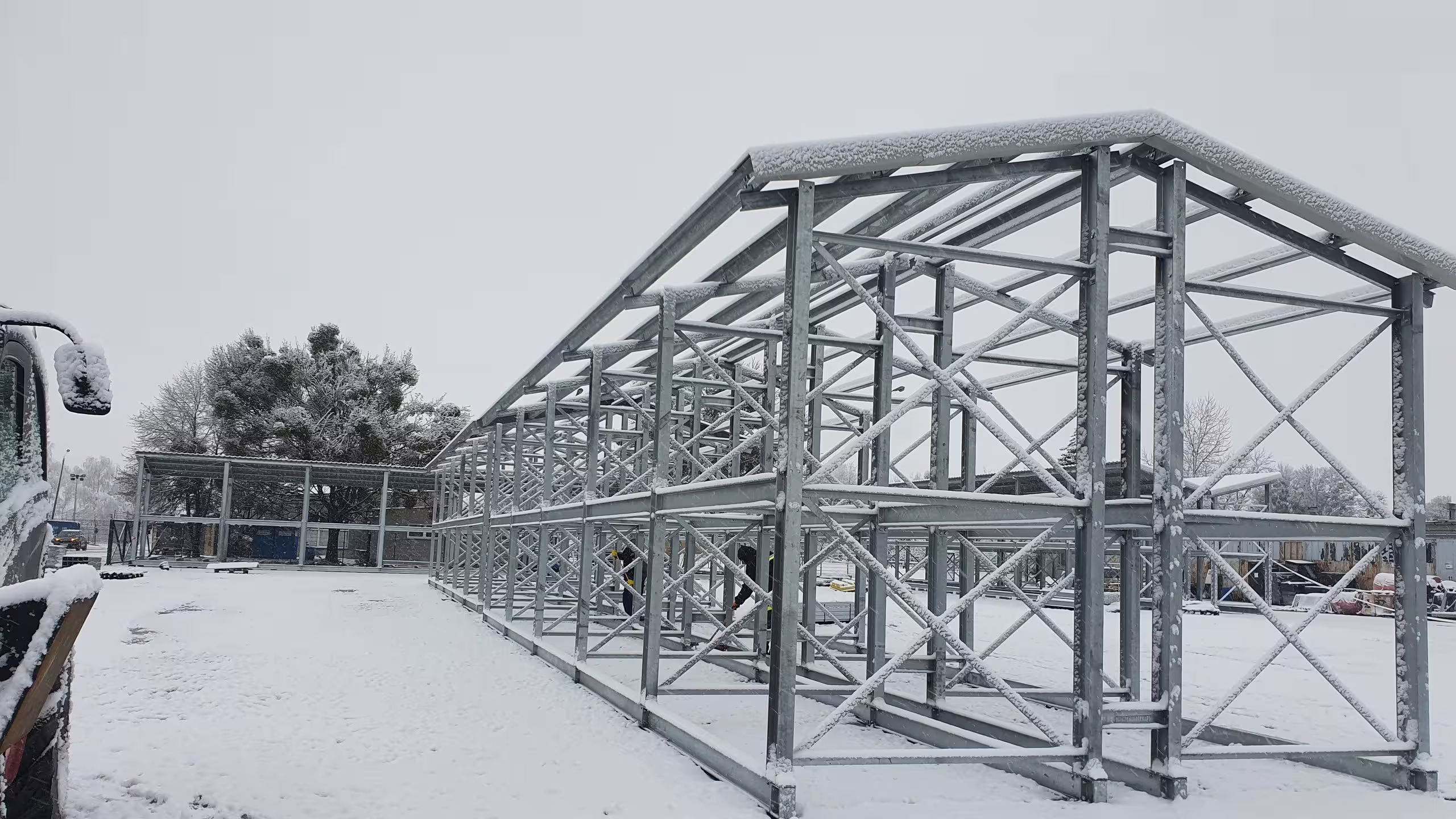 A galvanised CargoRack portal frame standing in a snow-covered yard with its lattice roof trusses and purlins erected and a layer of snow lying across them, a longer roofed run continuing behind it