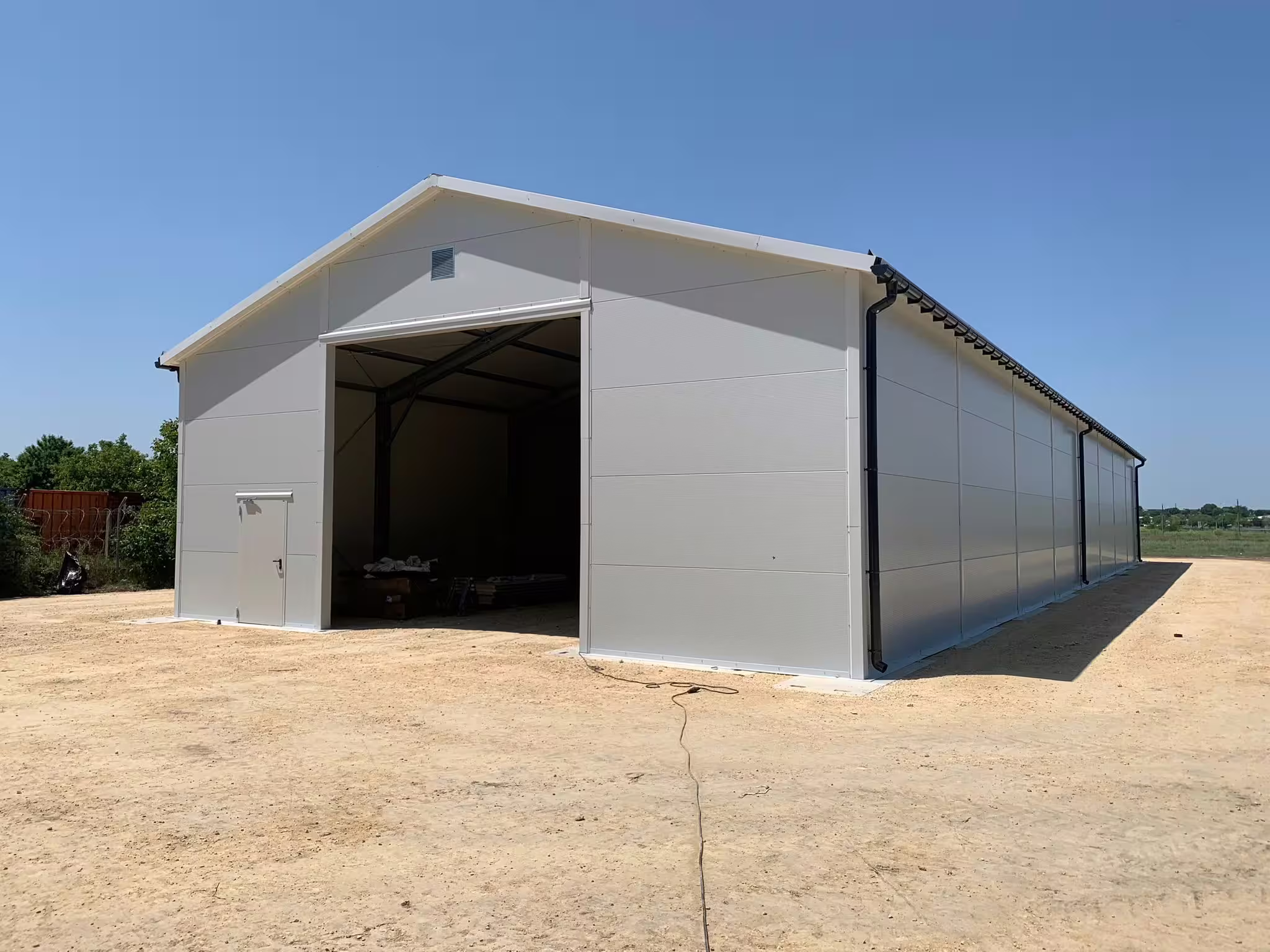 A DMTSS shelter on a sand compound with its full-width gable end open, showing an unobstructed internal span with material stacked along one side and no fixed fit-out