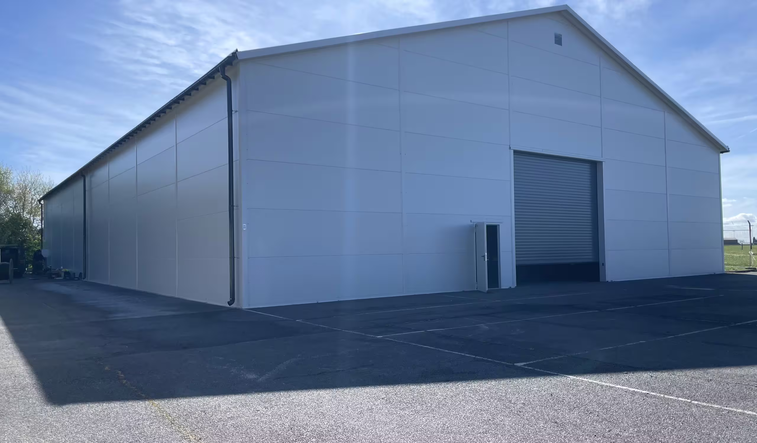 A white insulated-panel DMTSS shelter seen from the corner on a new asphalt apron under a blue sky, its gable end carrying a roller-shutter door and a personnel door, the long flank running away to the left