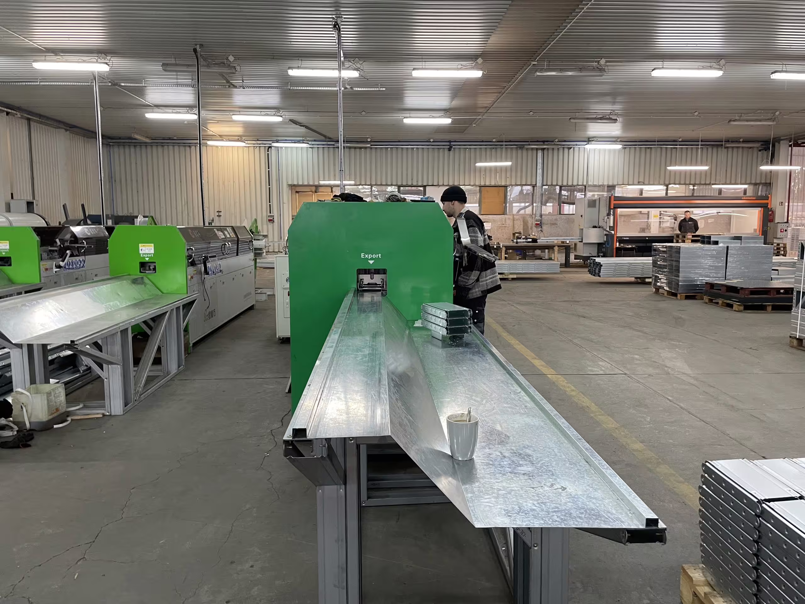 A long galvanised steel section running out of a green roll-forming line onto the outfeed bench, with further forming machinery down the bay