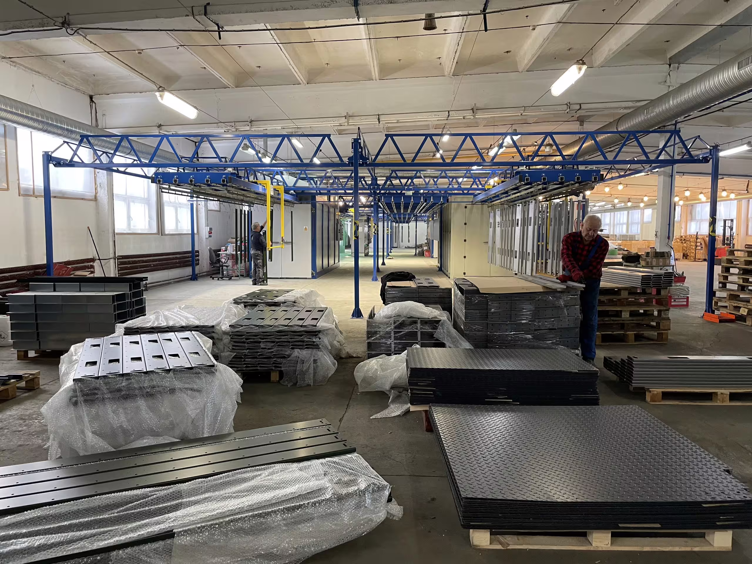 A factory bay with finished deck panels and wrapped sub-assemblies palletised down both sides under an overhead gantry, ready for dispatch