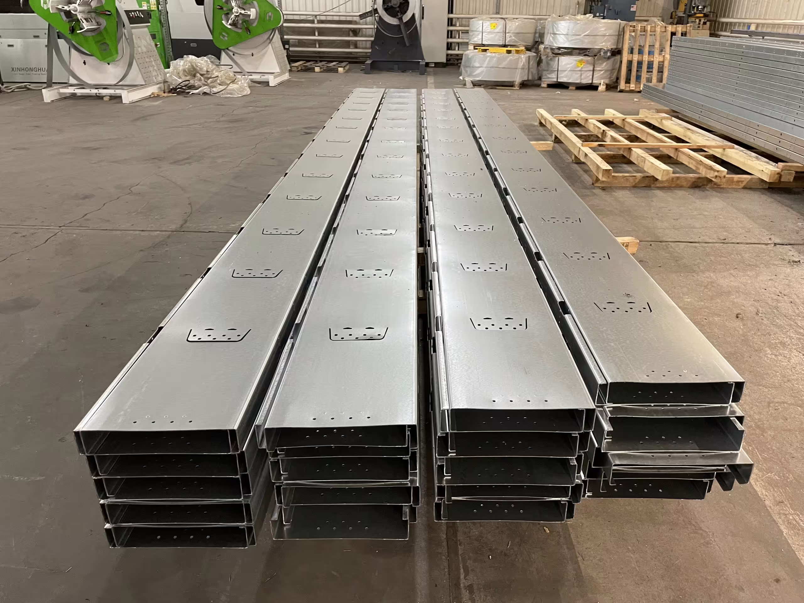 Rows of long galvanised steel sections laid out on a factory floor, punched and pressed ready for assembly, with pallets and further stock behind