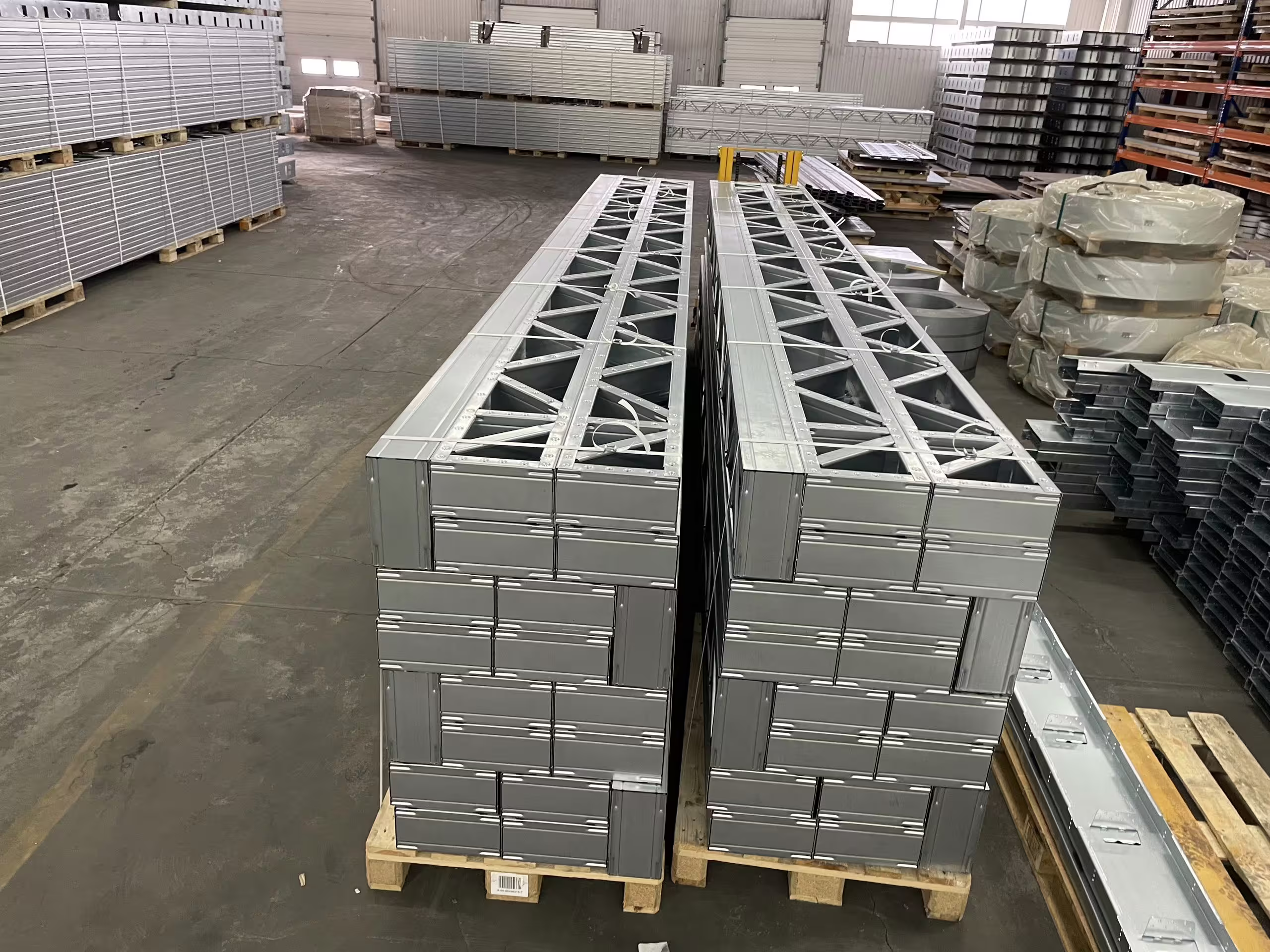 Stacks of galvanised lattice frames banded onto pallets in the BANAIR factory, with racked panels and further pallets behind them