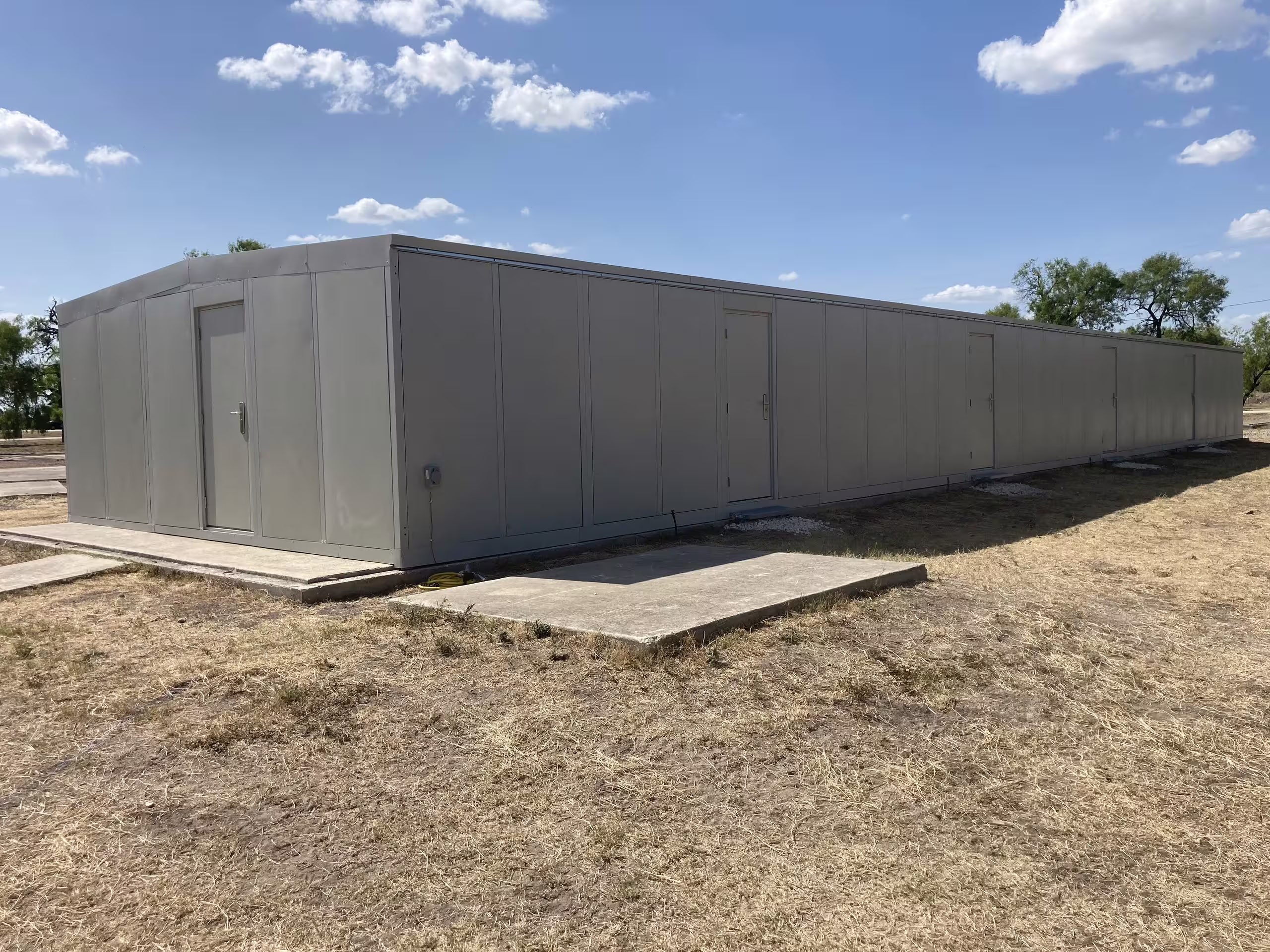 A long single-storey modular gas chamber trainer on dry open ground, its pale panel walls running away from a personnel door at the near end, concrete access pads at the thresholds