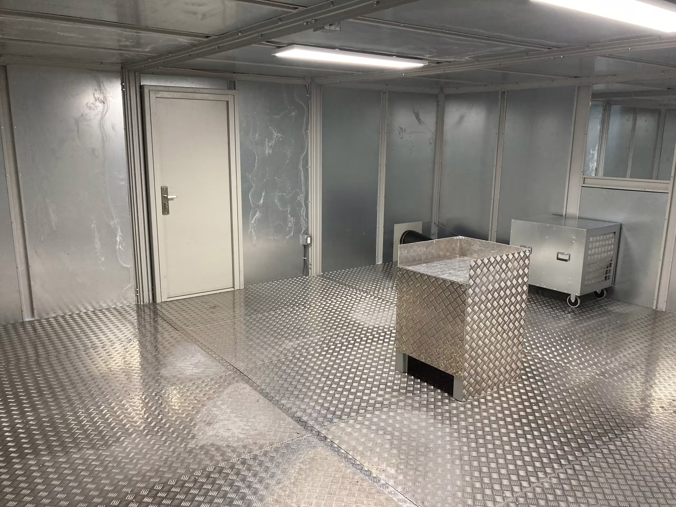 Inside the gas chamber trainer: a stainless burner pedestal standing on a chequer-plate floor, a wheeled cabinet behind it, a personnel door in the far wall and ceiling lighting running the length of the bay