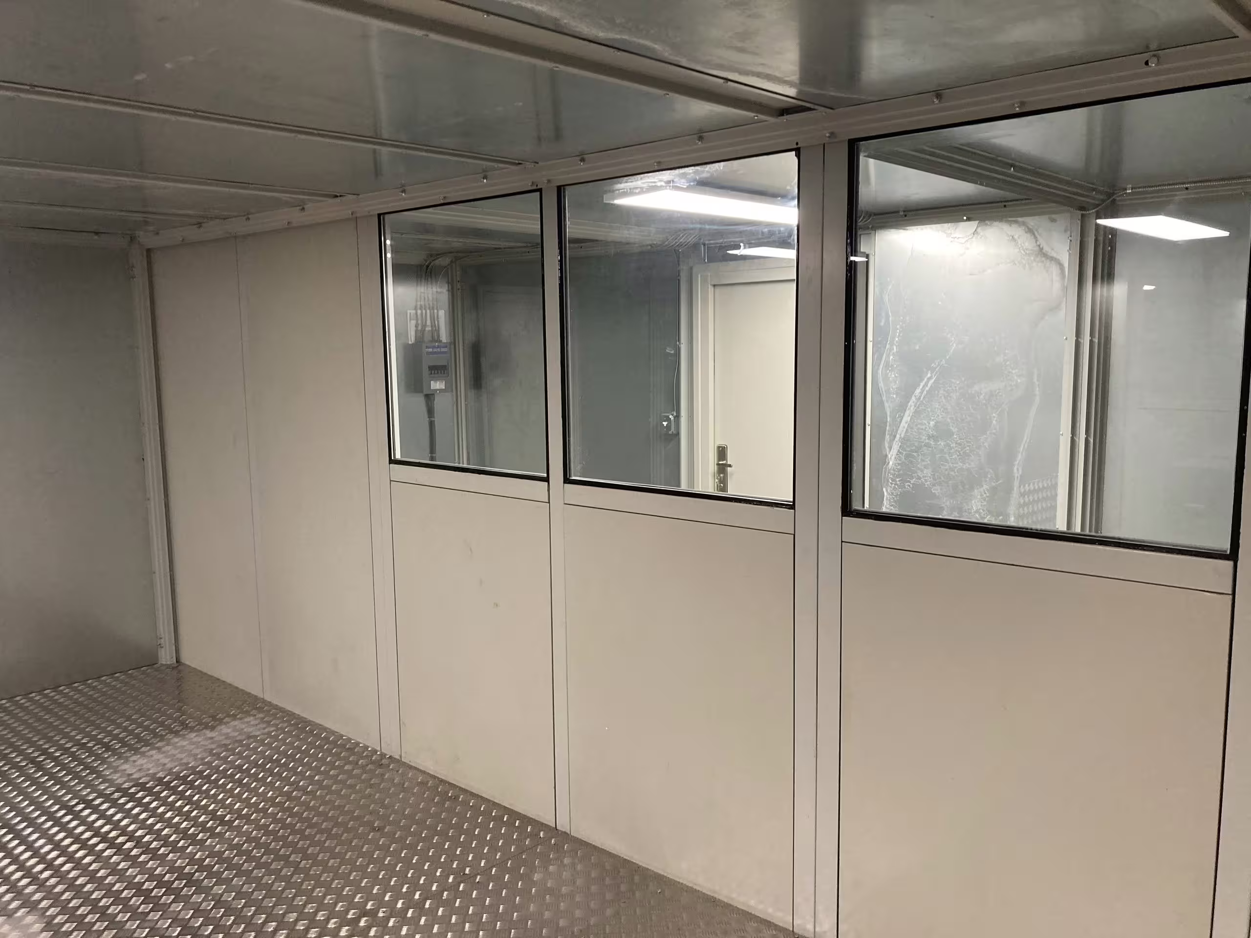 A run of large observation windows set into the chamber's internal partition, looking through to the lit space beyond, with a chequer-plate floor in the foreground