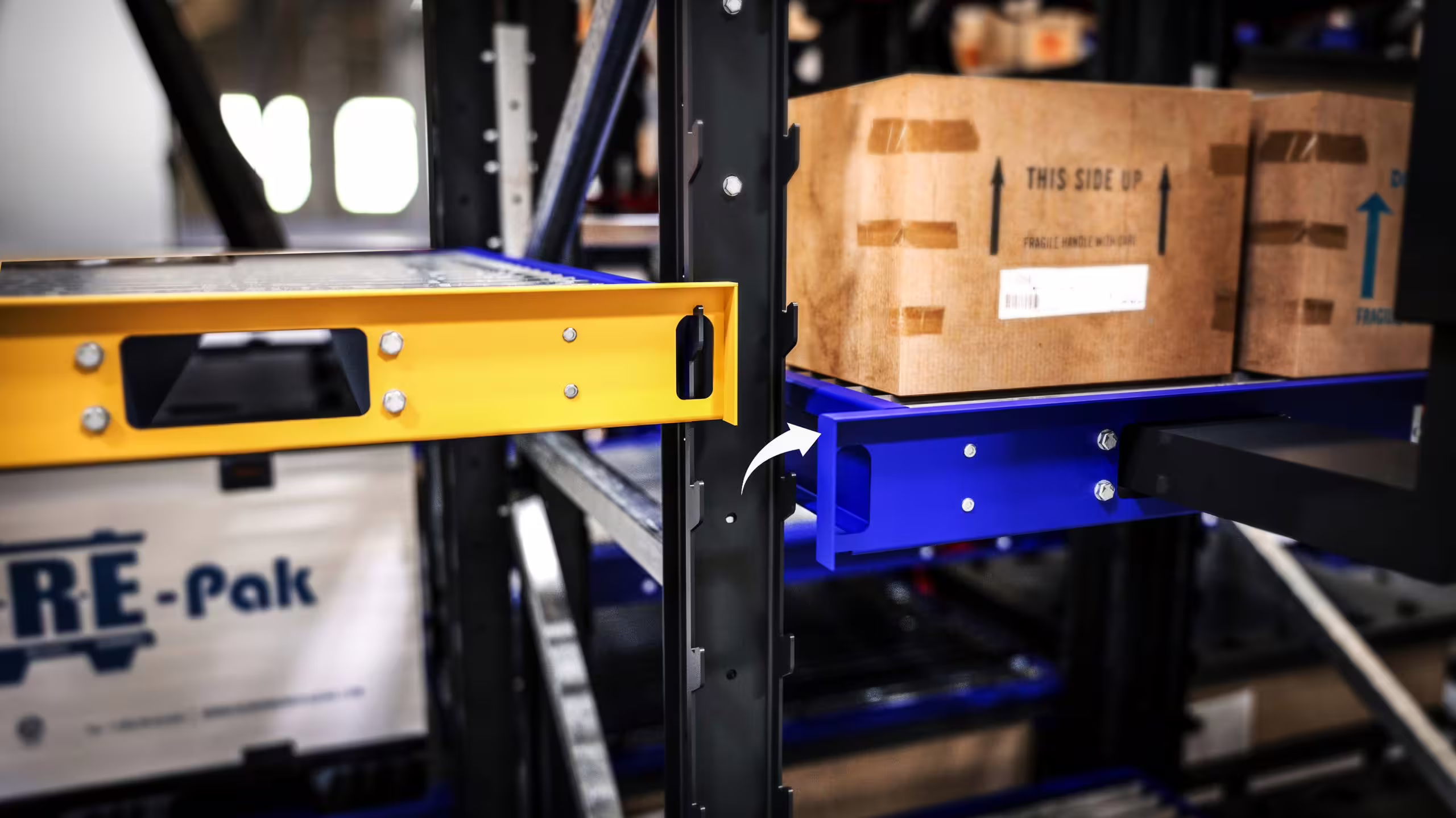 Close view of two IARS beams at an upright: a yellow beam lifted clear of the punched column with its hook disengaged, and a blue beam seated in the slots below it carrying a palletised carton