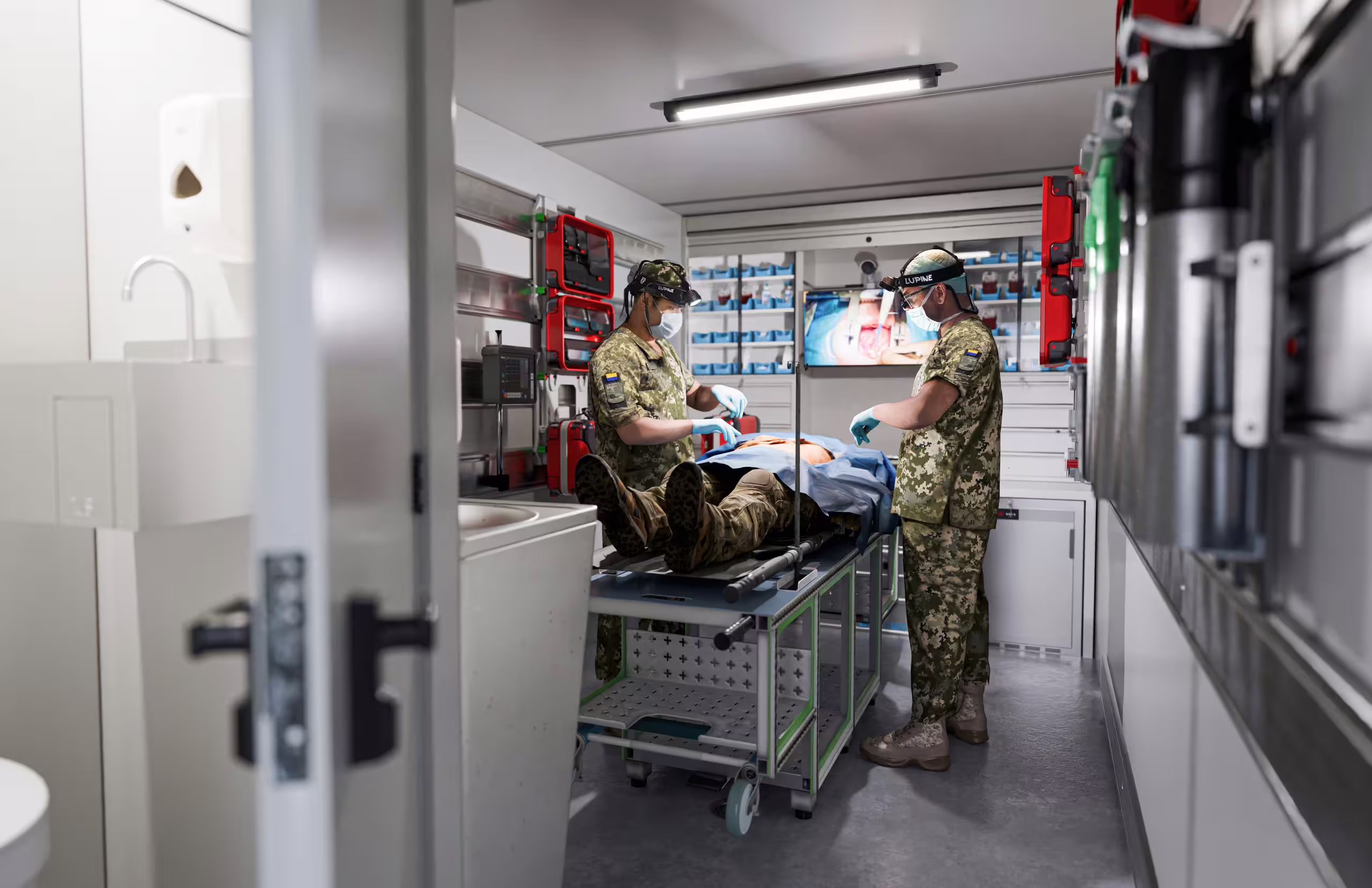 Two medics work over a patient on a trolley inside the fitted clinic, with supply shelving and a wall-mounted screen behind them.