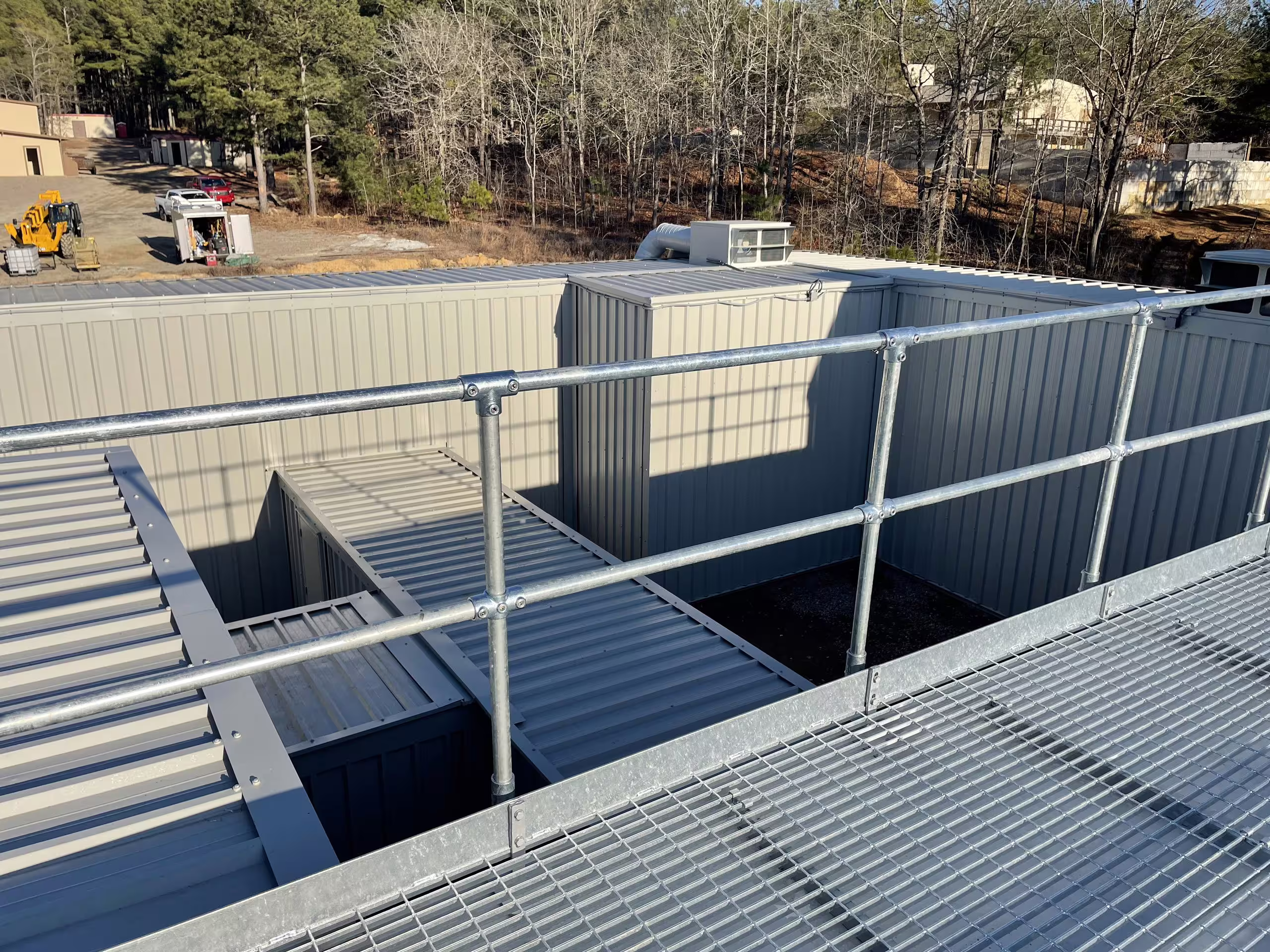 A galvanised mesh walkway with handrails running across the rooftops of a MOTC urban complex, open stairwells dropping into the structure below and ribbed-steel module walls on either side