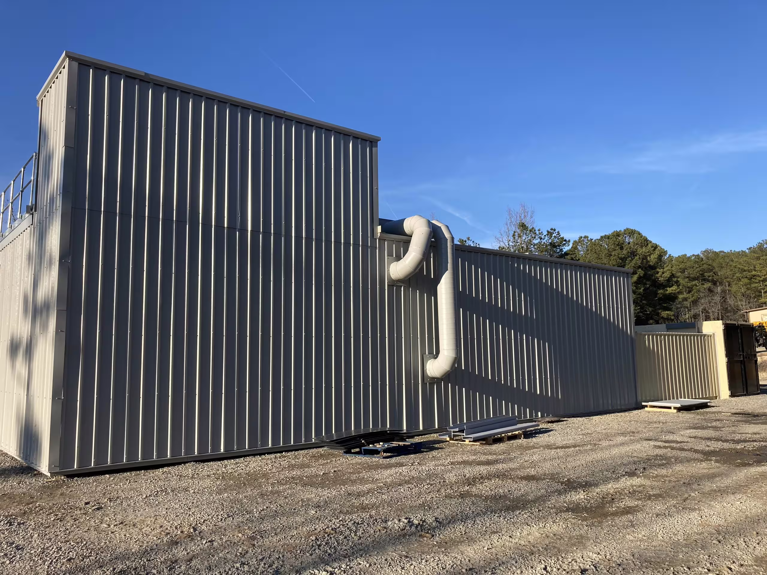 The flank of a MOTC urban block under a clear sky: ribbed steel panels rising two storeys to a stepped upper level, external ventilation ducting curving down the wall, gravel hardstanding at its base