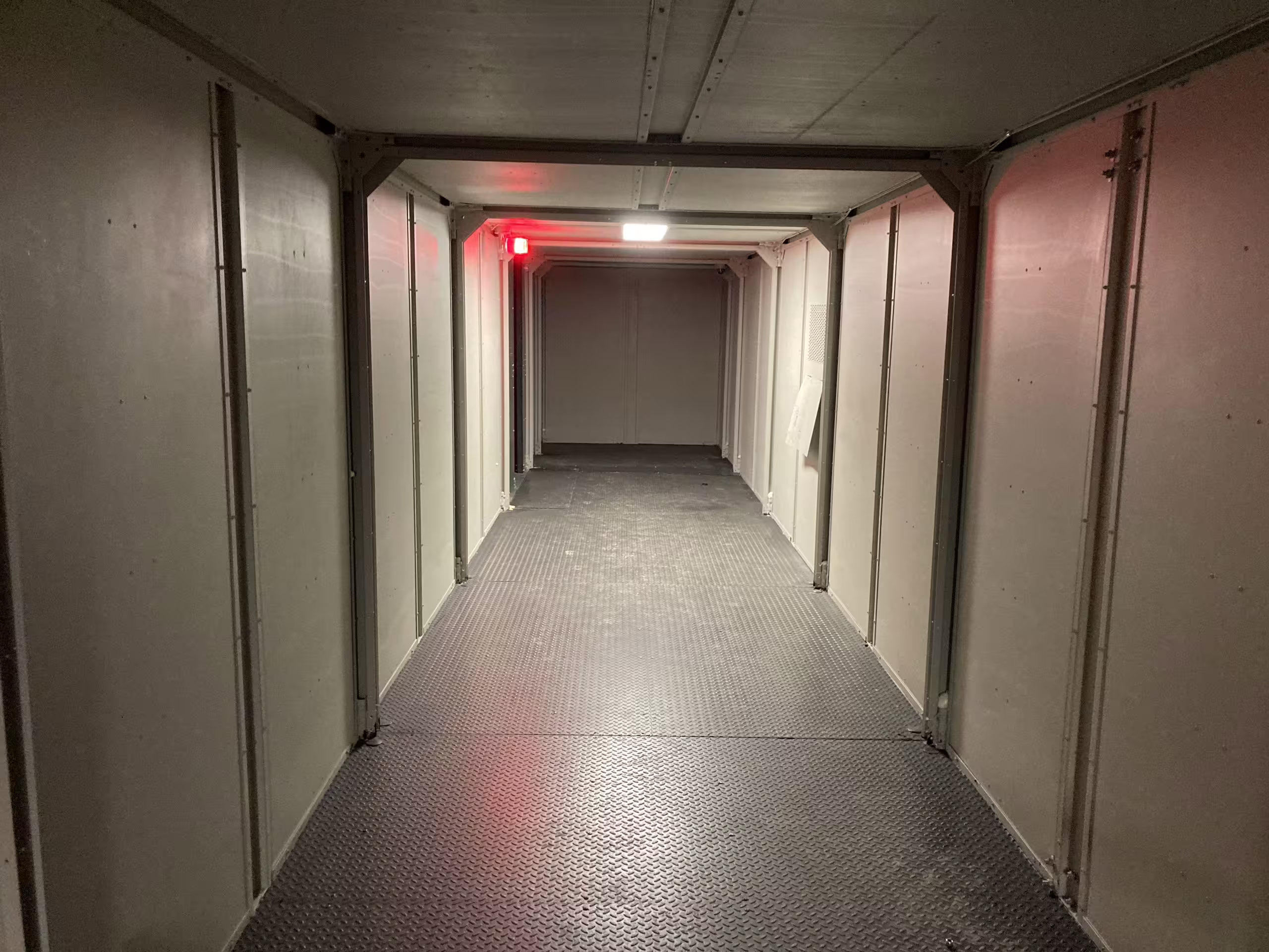 A corridor running the length of a MOTC module: panel walls and door openings on both sides, chequer-plate floor, ceiling lighting overhead and a red lamp burning at the far end