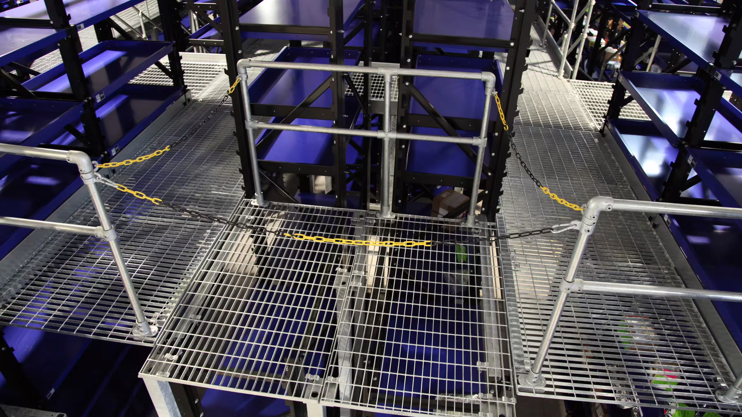 Looking down onto a RackMezz mezzanine walkway: galvanised open-mesh grating panels, tubular handrails on both sides and yellow safety chains slung across a transfer opening, with blue shelf units below on either side