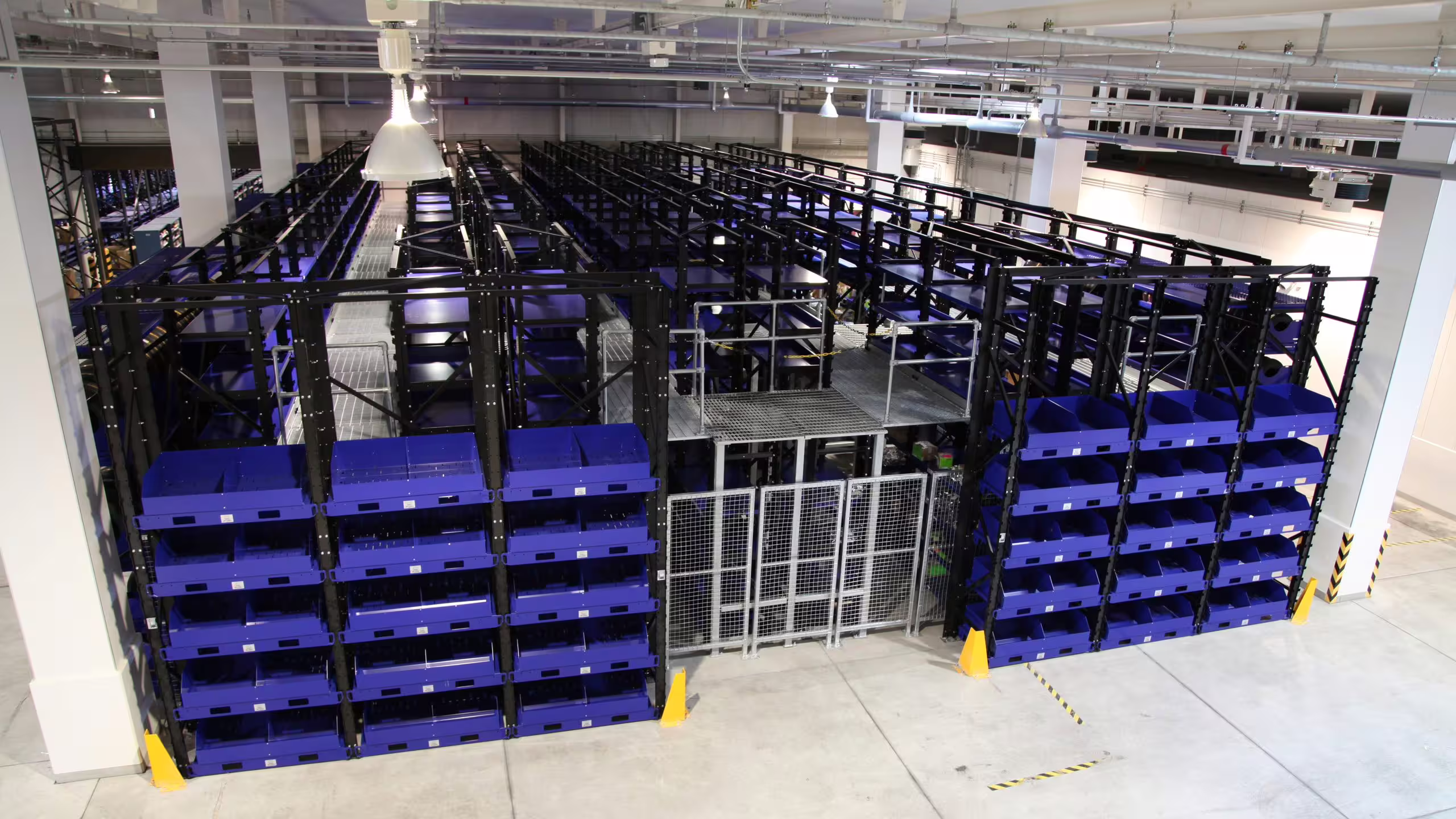 An elevated view across a complete RackMezz installation: an open mezzanine deck of galvanised grating with tubular handrails carried over a lower level of blue open-front bins, a mesh security cage beneath the deck and yellow floor guards at the base