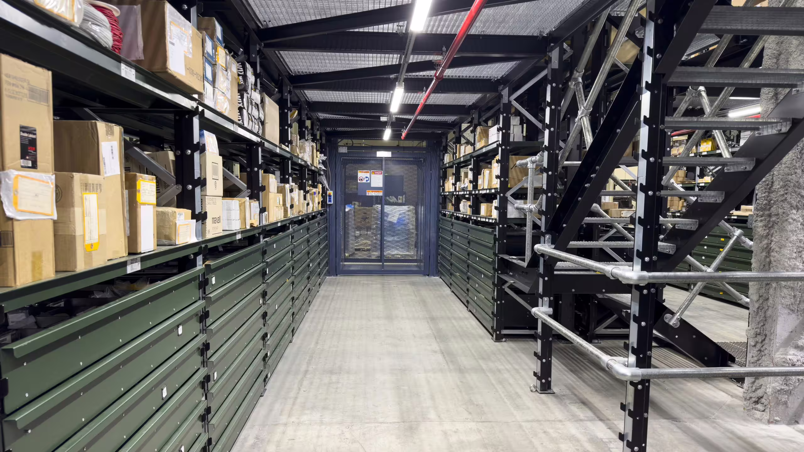 A warehouse aisle lined with green RackMezz drawer units, with a bolted steel staircase and tubular handrail climbing to the mezzanine deck on the right