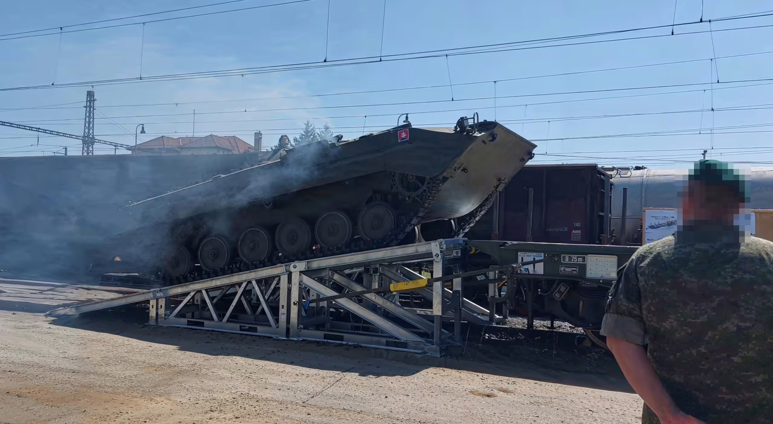 A tracked armoured vehicle driving down a galvanised ramp from a rail flatcar onto hardstanding, exhaust smoke behind it and a marshal standing clear at the right