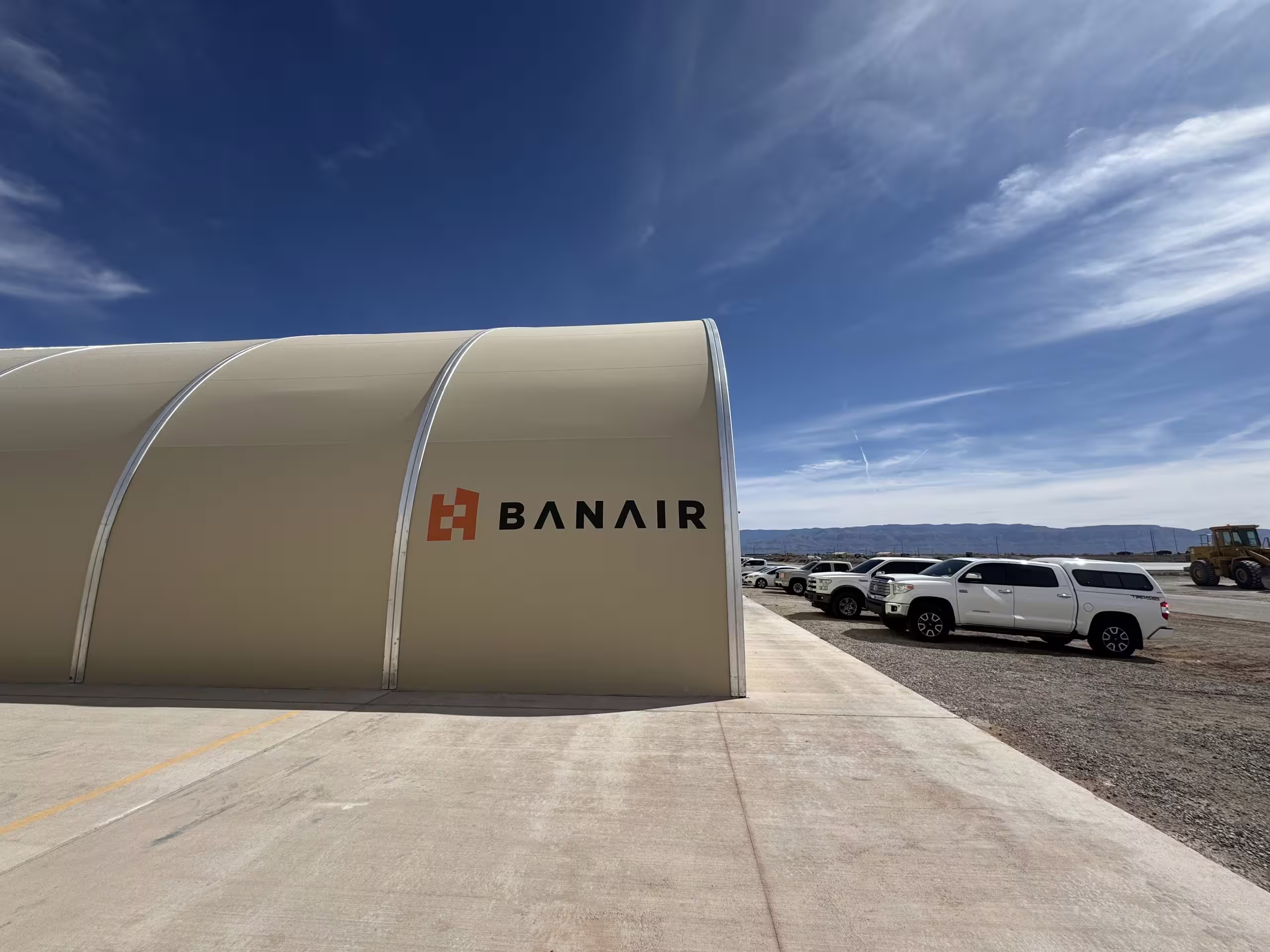 A tensioned-membrane Sprung shelter on hardstanding under a blue sky, its curved white roof carrying the BANAIR wordmark, vehicles parked alongside