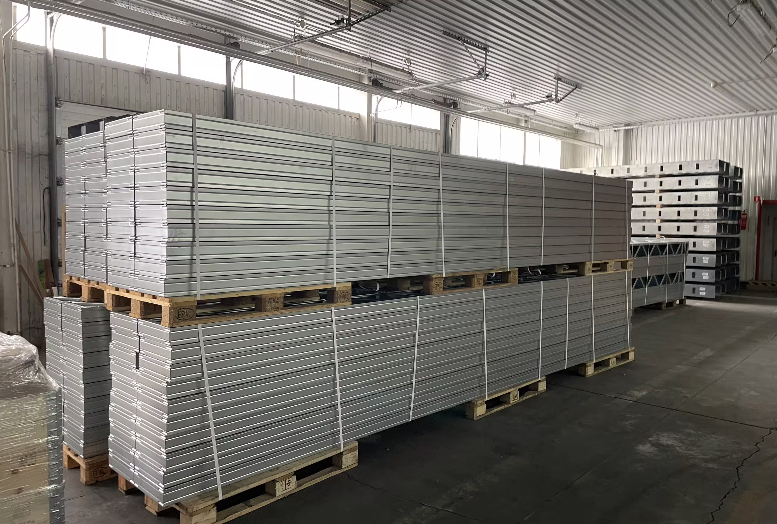 Inside BANAIR's factory: a long run of insulated wall panels stacked on timber pallets down the length of the bay, with racked steel sections beyond and a swept concrete floor