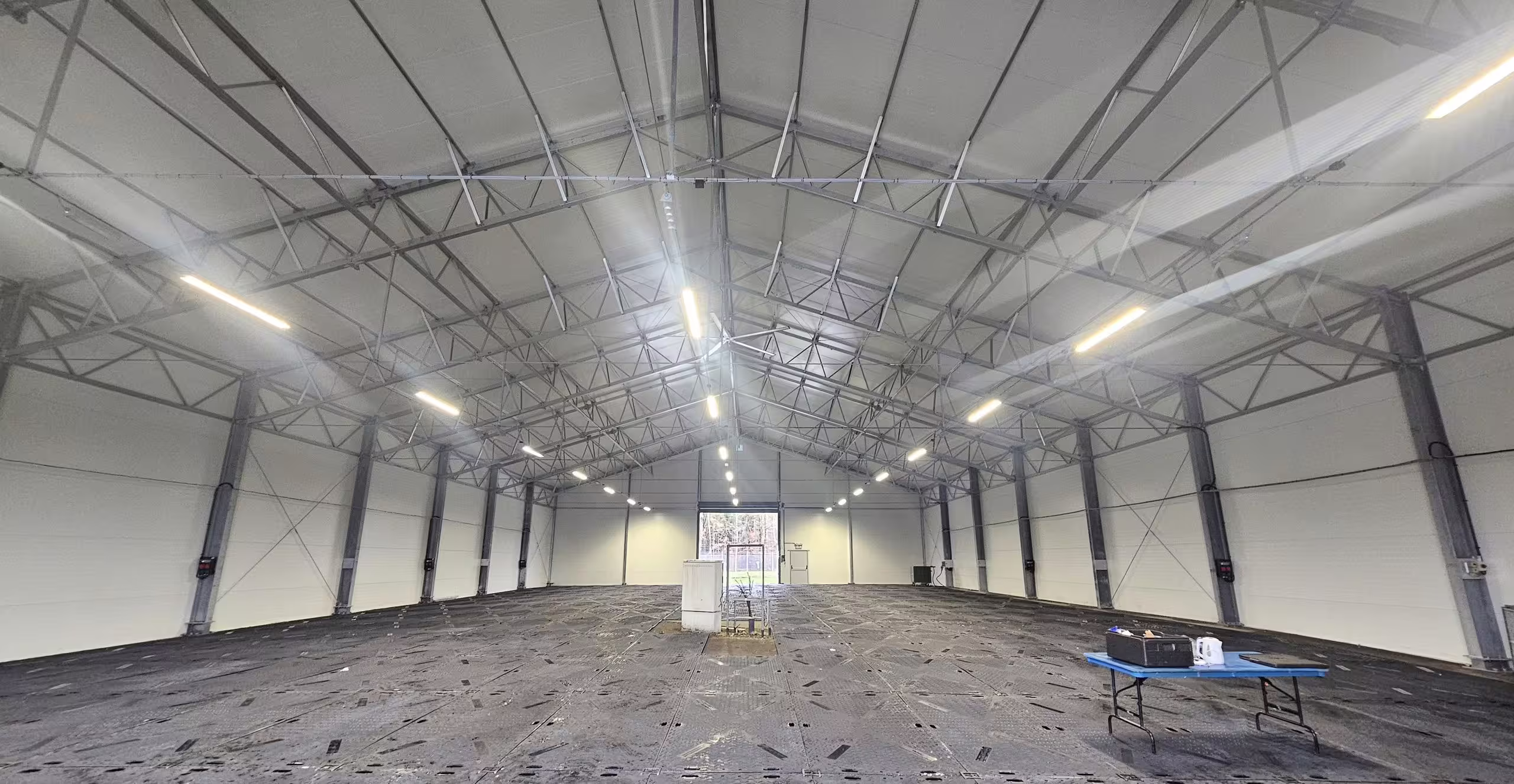 The interior of a DMTSS shelter looking down its length: a clear unobstructed span under a lined roof with continuous lighting runs, roller-shutter doors in the end wall and a bare floor