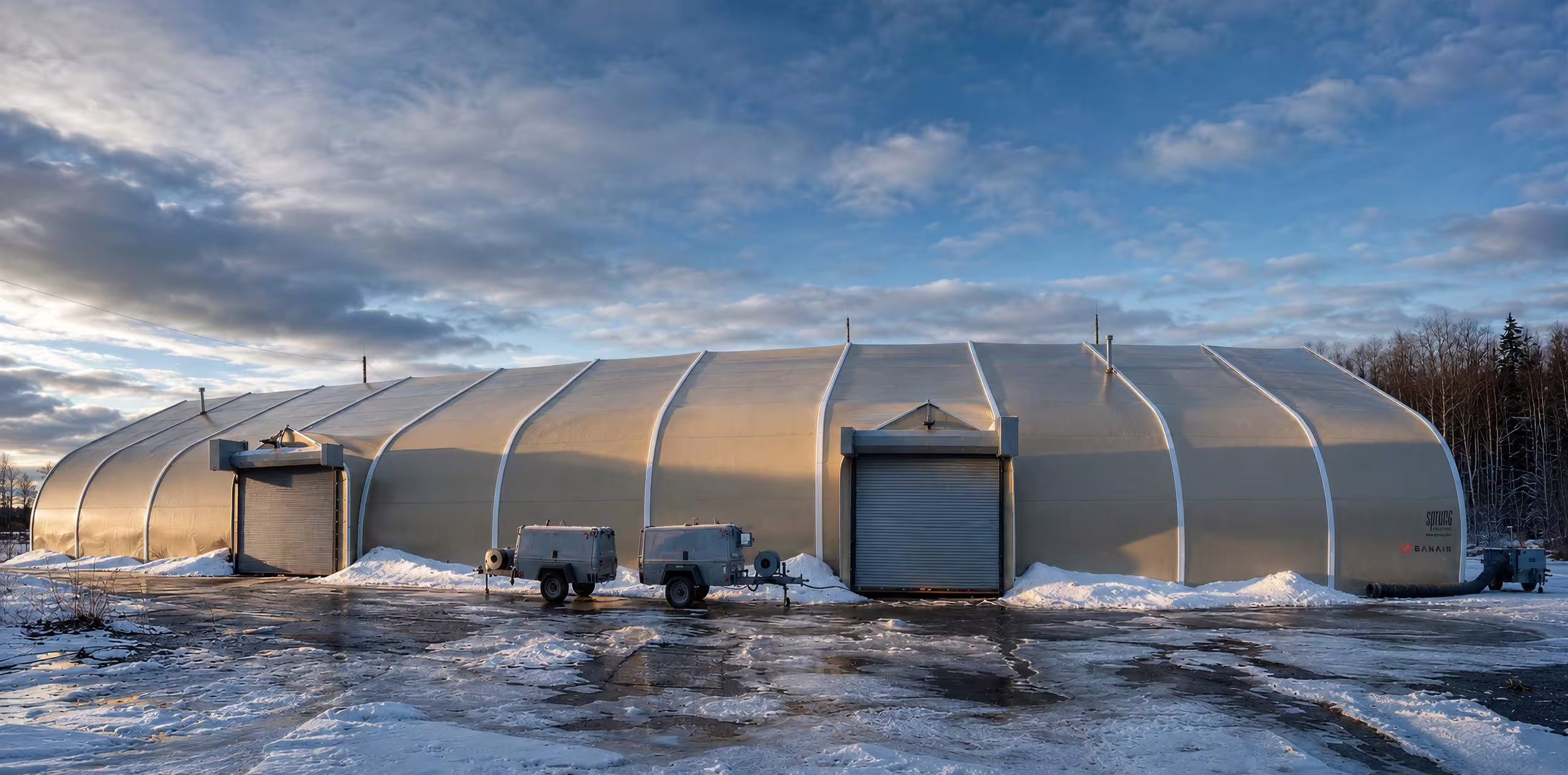 A long tan fabric-membrane Sprung shelter photographed along its flank in deep snow under a broken sky, with two roller-shutter doors and service units set against the base