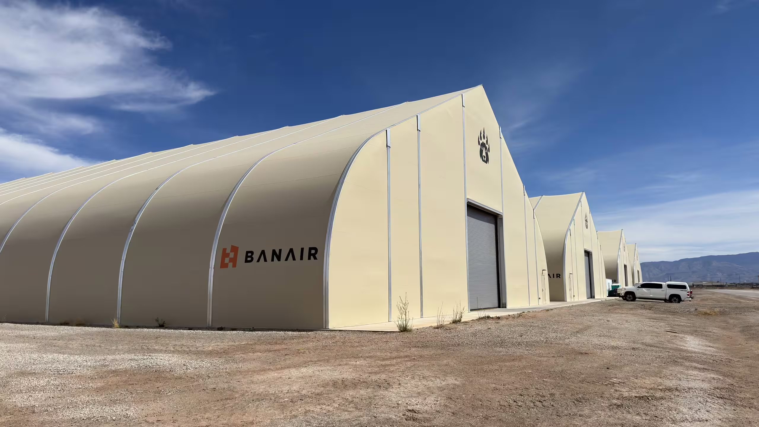 Three tan fabric-membrane Sprung shelters standing in a row on desert hardstanding under a deep blue sky, the nearest carrying the BANAIR wordmark across its flank and a roller-shutter door in its gable, a mountain range on the horizon