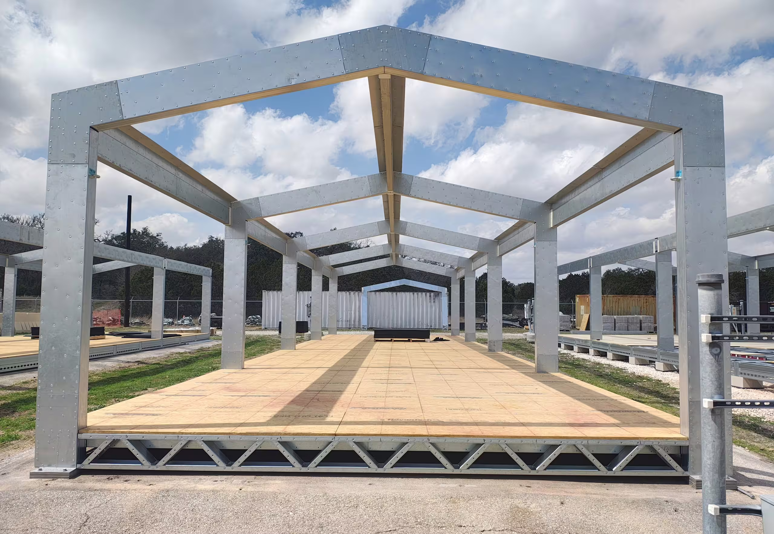 A bolted shelter frame standing square-on partway through erection: galvanised portal frames and ridge beams over a board deck carried on lattice floor joists, with further frames standing either side under a broken sky