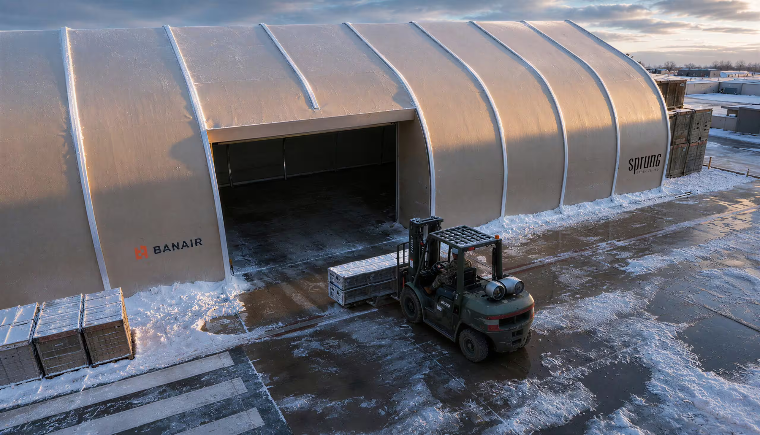Elevated view at dusk of a Sprung shelter with its end door open, carrying the BANAIR wordmark on one bay and the Sprung Structures wordmark on another, as a rough-terrain forklift carries a palletised load towards the opening across a wet, part-cleared apron with crates stacked alongside
