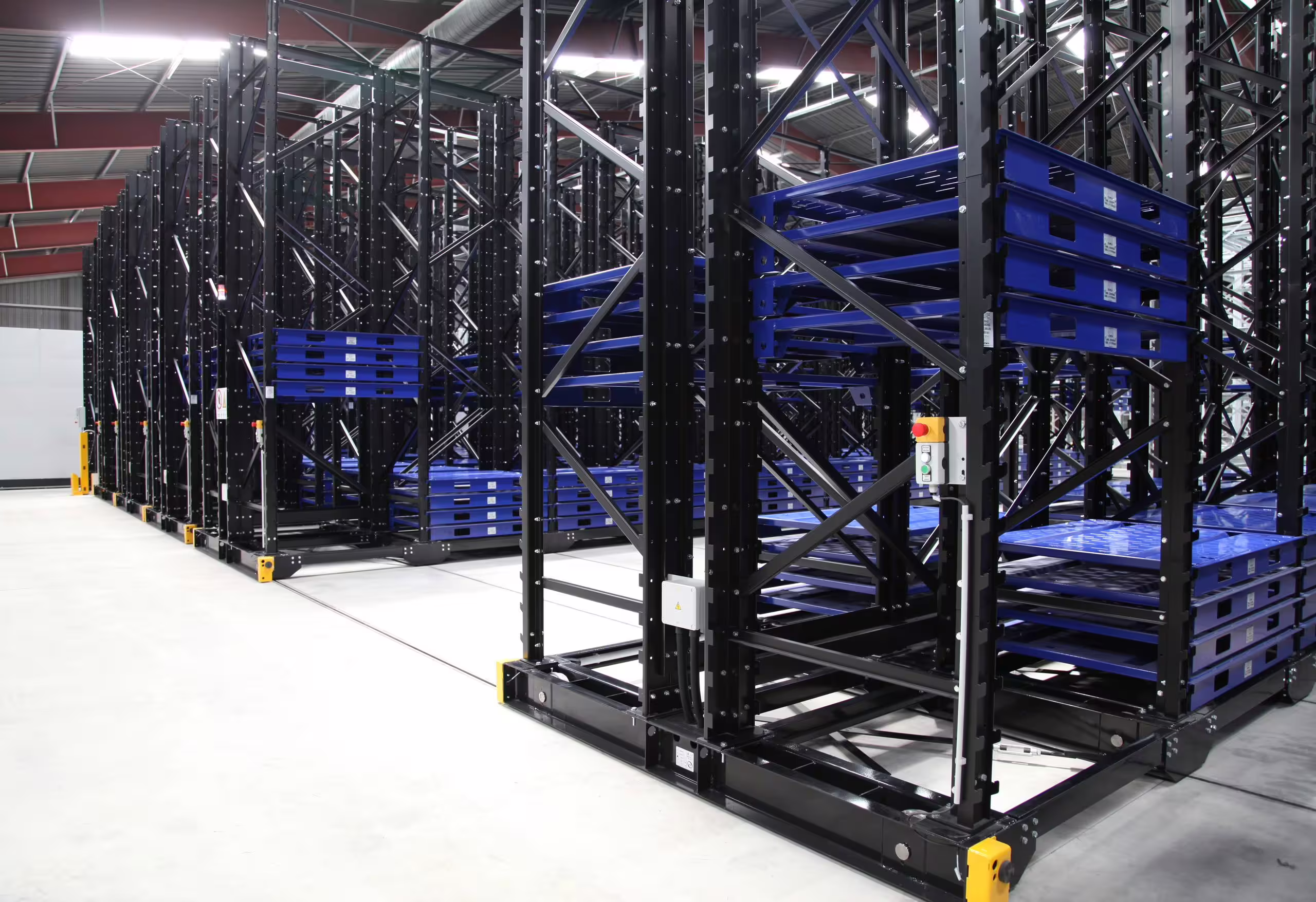 Inside a storage hall: several bays of black mobile racking standing on floor rails, each bay carrying stacked blue steel trays, with push-button control stations and red emergency stops on the end frames and yellow guards at the rail ends