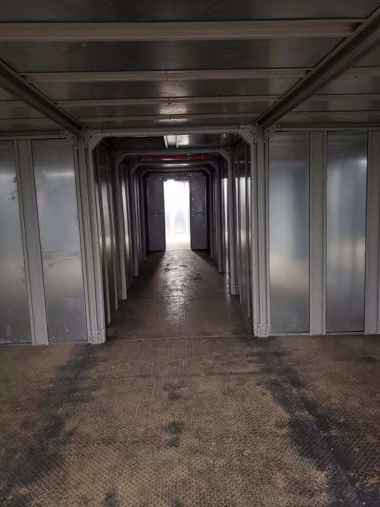 Looking down the inside of a subterranean trainer tunnel: ribbed metal walls and ceiling closing in on both sides, a bare floor underfoot and daylight at the far opening