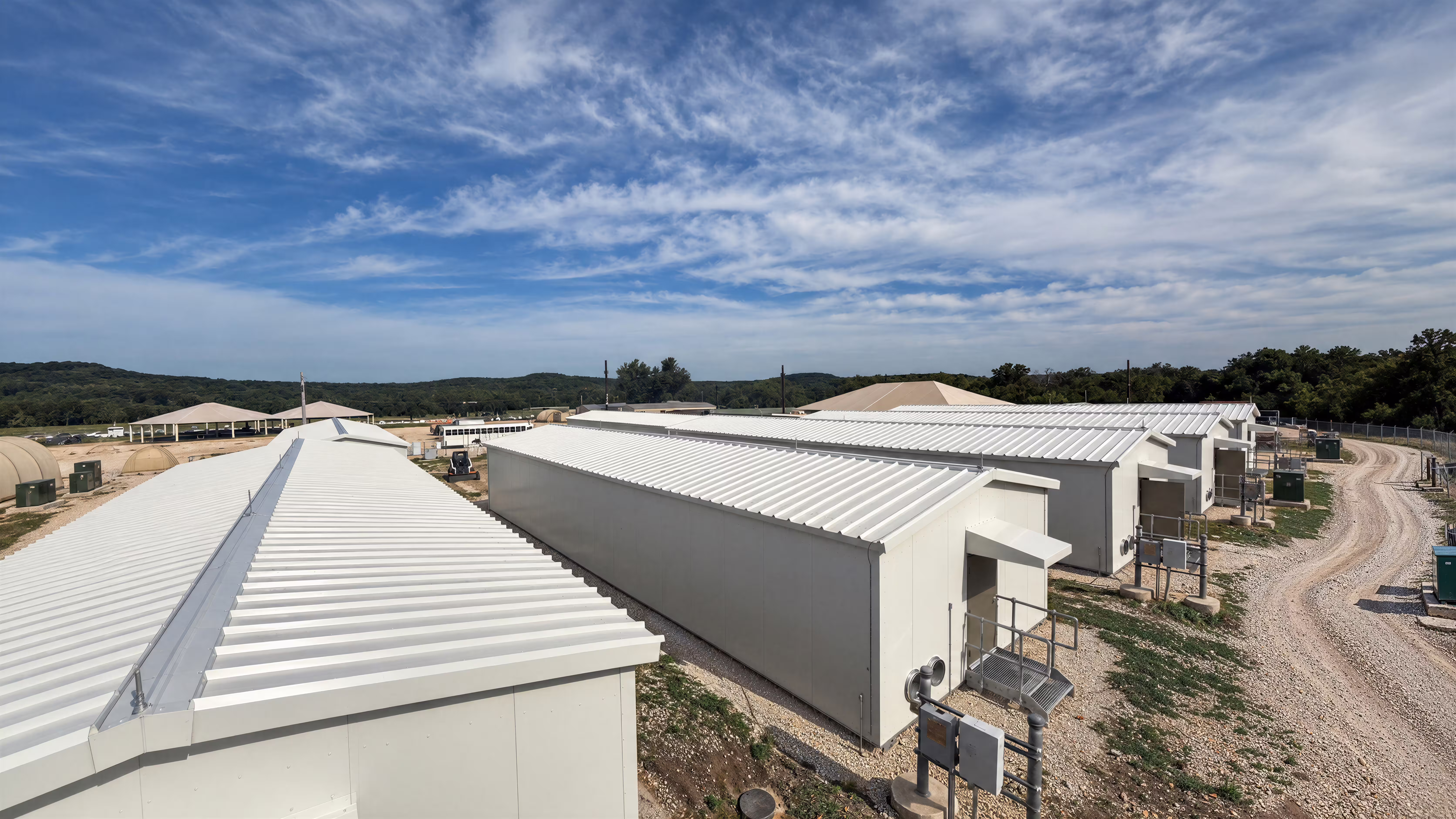 Aerial view along a row of Tactical Shelter buildings on hardstanding: white standing-seam roofs and pale insulated wall panels, personnel doors and services panels down the near elevation, a gravel access road to the right and wooded hills on the horizon