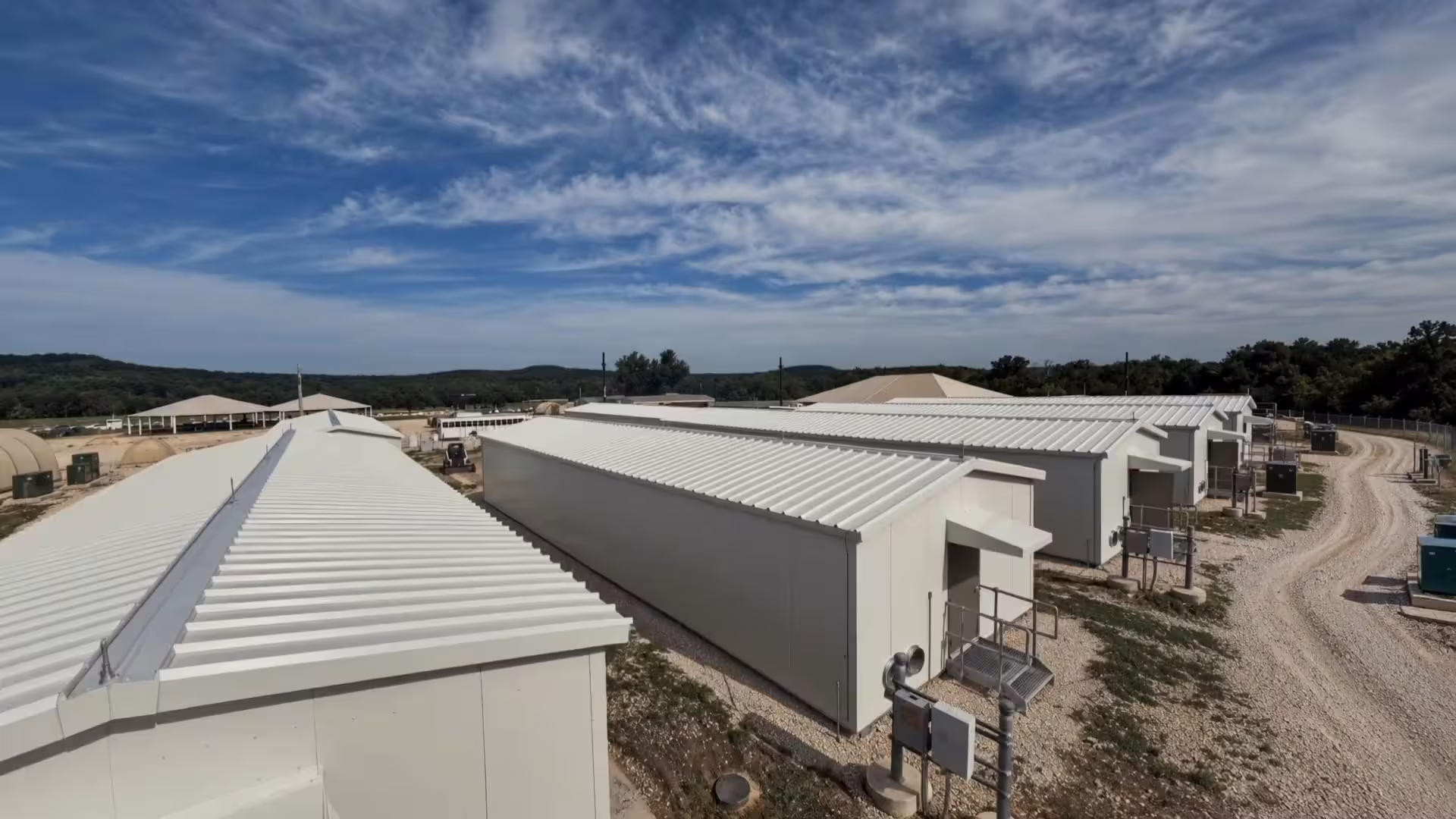 An elevated view along a row of white hard-wall tactical shelters on gravel, their ribbed roofs running into the distance under a wide blue sky streaked with high cloud, steel entry stairs and electrical panels along their sides, tan shade canopies and arched fabric shelters beyond, and a gravel track curving away to a tree line